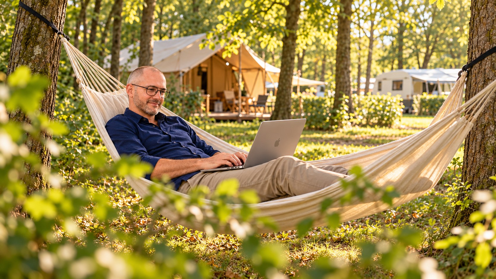 A man wearing glasses works on a laptop in a hammock strung between two trees at a campsite in golden evening light